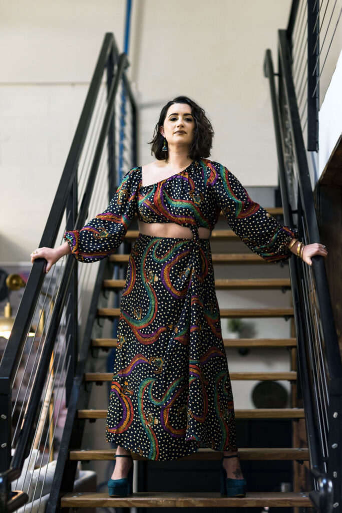 Portrait of author Victoria Margaux Nielsen. Margaux presents as a white woman with shoulder-length dark wavy hair, deep crimson lipstick, and a long two piece black dress with white polka dots and red and green swirly stripes. She is standing on a staircase in front of a white wall.